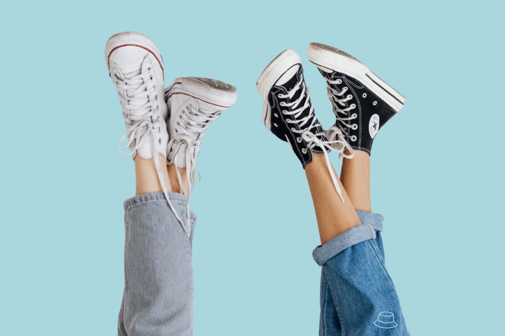 Two pairs of legs in jeans raised up, wearing white and black high-top sneakers against a blue background.